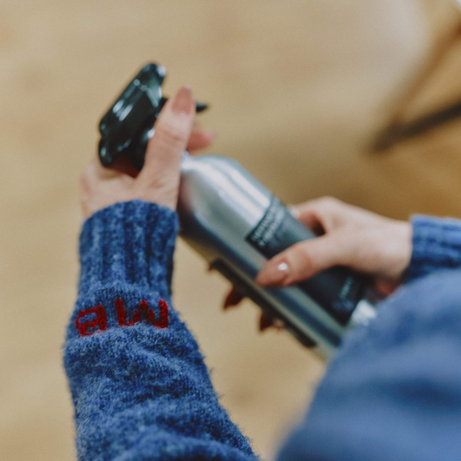 A close-up lifestyle shot of a Morgan Bagshaw, founder of Lavender and Lemon, holding a silver and black All Purpose Cleaner spray bottle, wearing a blue knit cardigan by Genevieve Sweeney with her red embroidered initials on the cuff.