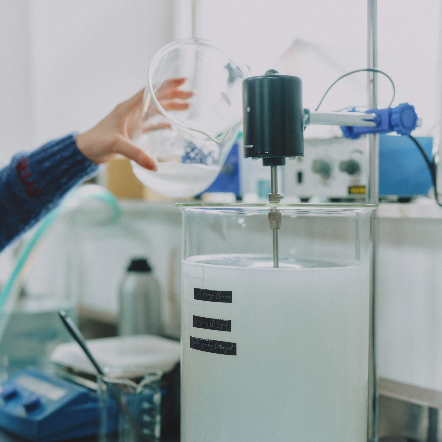 Morgan Bagshaw, founder of Lavender and Lemon, in a blue cardigan pours a clear liquid from a glass beaker into a large glass mixing vessel with an electric motorized stirrer, at her studio in Wales where she formulates a range of hand made, naturally derived cleaning products.