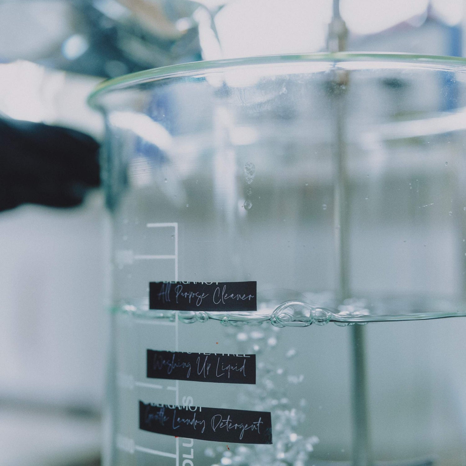 A clear glass laboratory beaker filled with water, showing measurement markings and labels for Lavender and Lemon household cleaners including All Purpose Cleaner, Washing Up Liquid, and Laundry Detergent. The background is a softly blurred, cool-toned indoor setting at her studio in Wales.