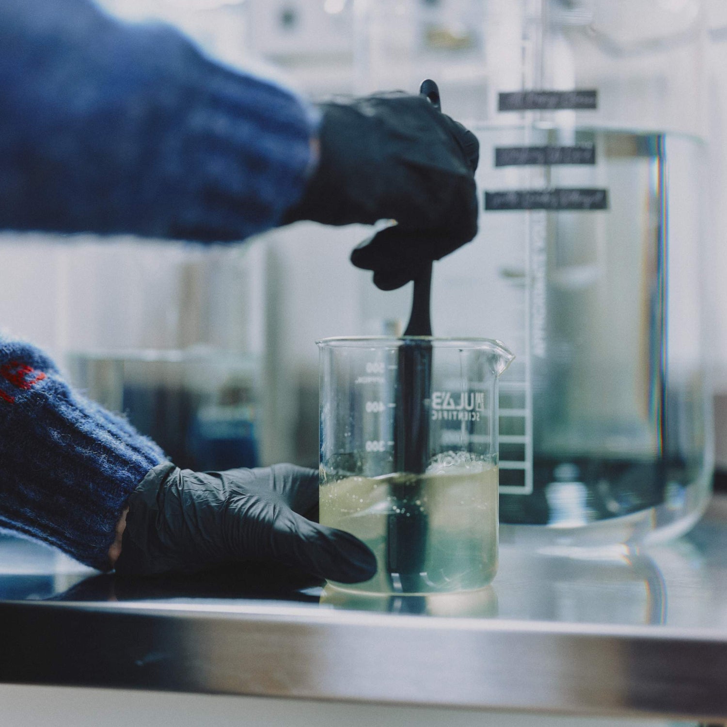 Morgan Bagshaw, founder of Lavender and Lemon, wearing black gloves stirs a liquid in a clear glass beaker on a stainless steel lab counter, with laboratory equipment blurred in the background.