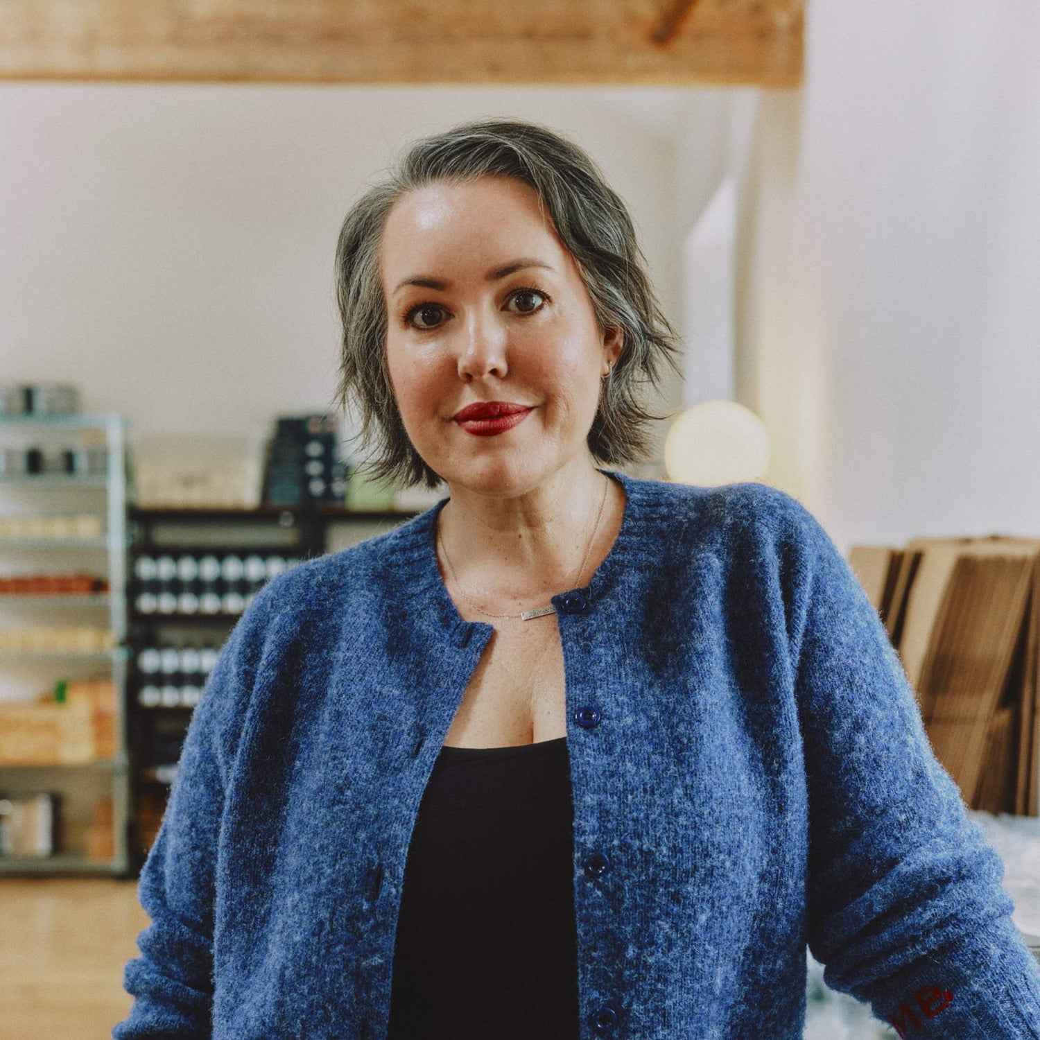 Morgan Bagshaw, founder of Lavender and Lemon, wearing a textured blue button-up cardigan by Genevieve Sweeney over a black top, standing in her studio in Wales with shelves of Lavender and Lemon products  in the background.