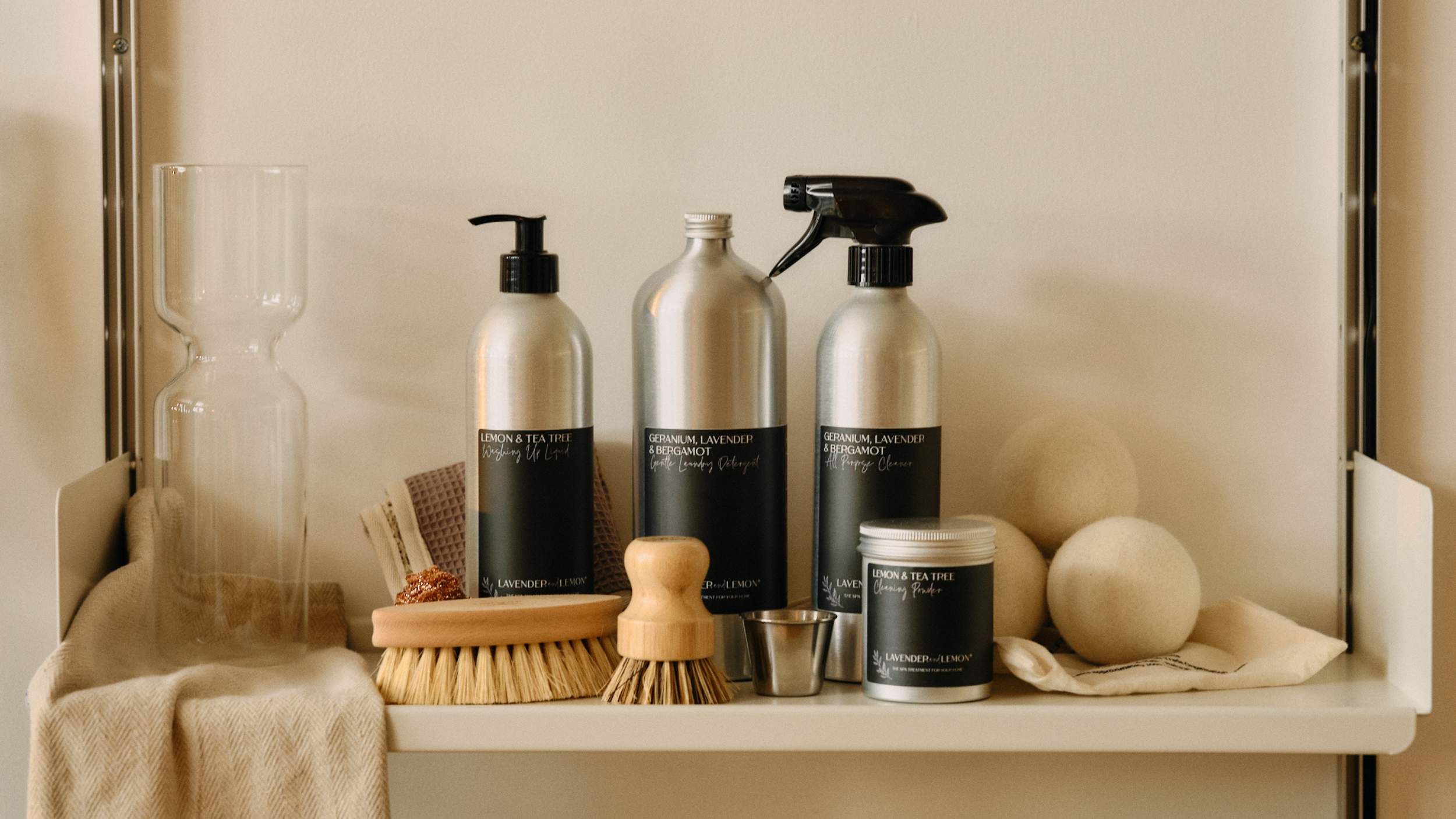 A curated collection of home cleaning products from Lavender and Lemon on a white shelf, including silver bottles with black labels, a spray bottle, a small tin, wooden-handled brushes, and natural fiber cloths, styled with neutral props against a warm beige background.