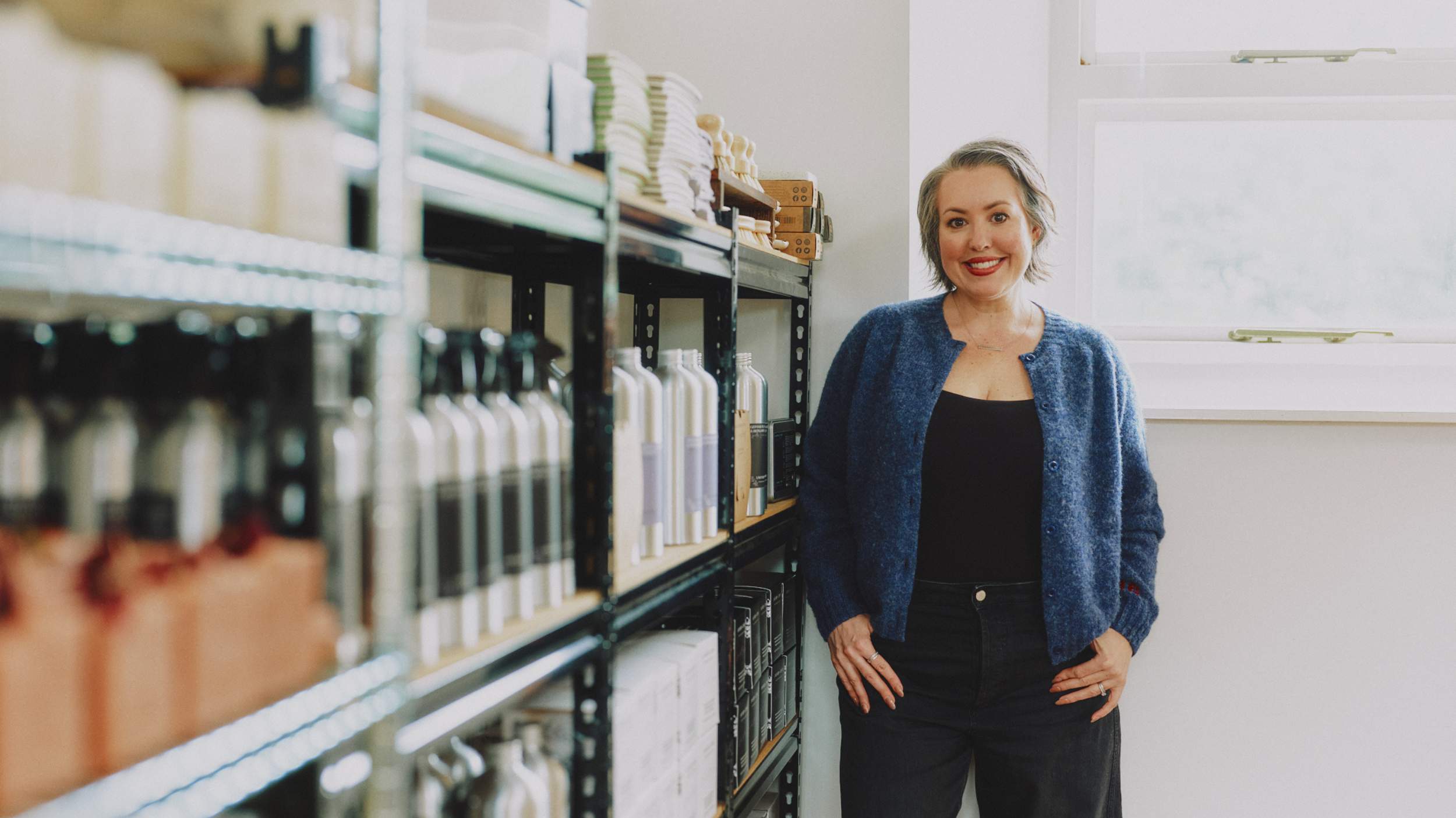 Morgan Bagshaw, Founder of Lavender and Lemon, stands smiling in a bright room next to shelves filled with Lavender and Lemon products, wearing a blue knit cardigan by Genevieve Sweeney over a black top and black jeans.